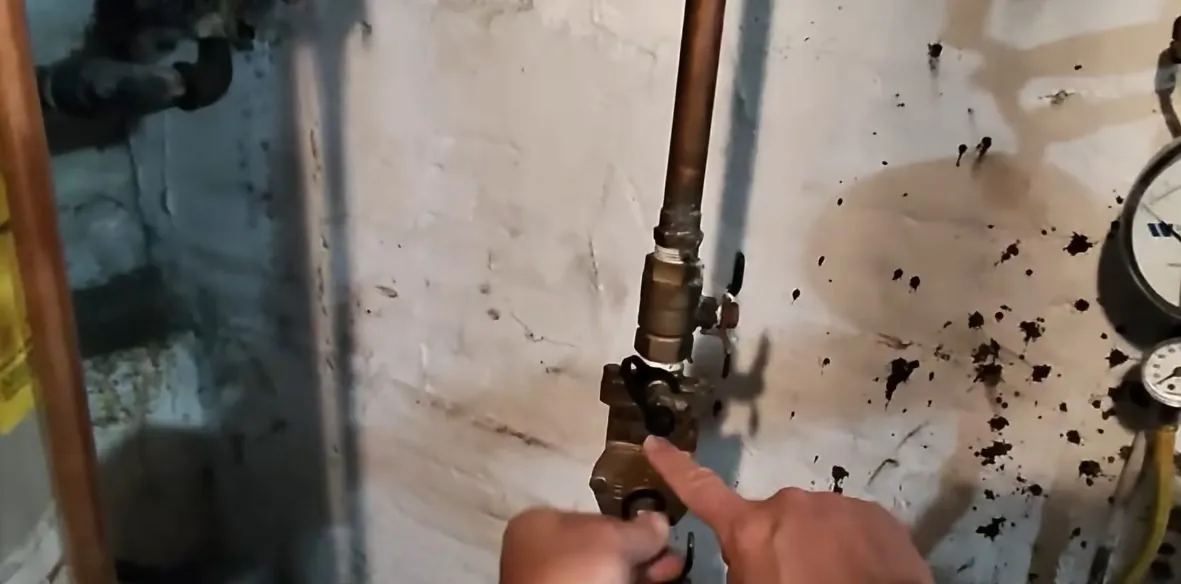 Hand pointing at a brass shutoff valve on a copper pipe against a basement wall