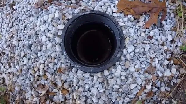 Open black cleanout pipe set in a bed of white gravel with a few fallen leaves